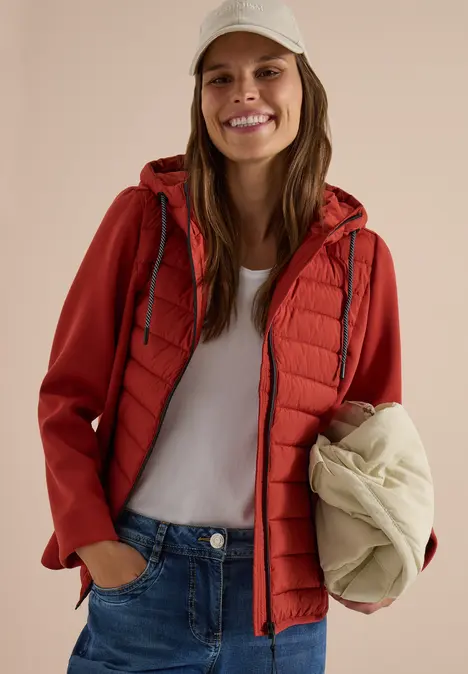 Frau in roter Jacke und weißem Shirt, lächelt, hält eine cremefarbene Weste in der Hand.
