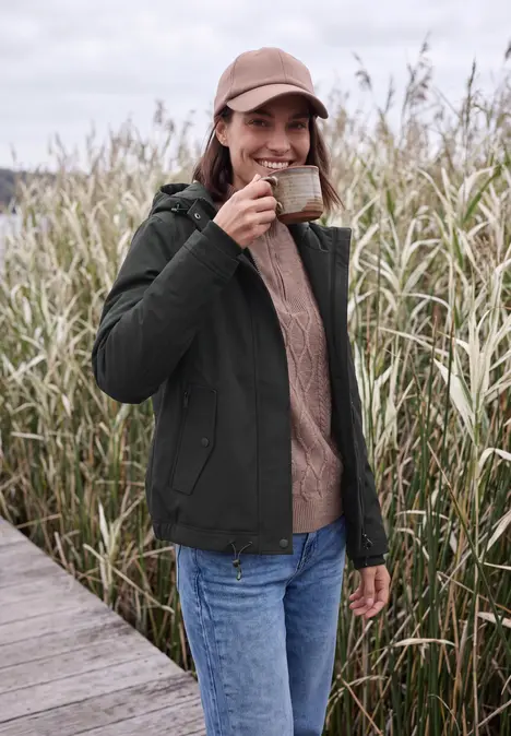 Eine Frau in Jacke, Jeans und Cap trinkt Kaffee aus einer Tasse auf einem Steg neben hohem Schilf.