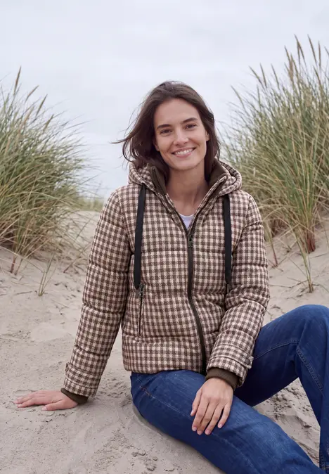 Junge Frau sitzt lächelnd in kariertem Mantel und Jeans auf Dünen mit Strandgras.