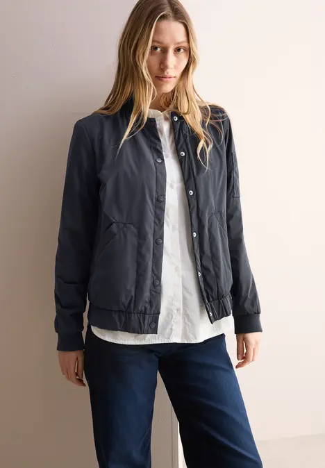 Frau mit langer Bluse, dunkelblauer Jacke und Jeans steht vor heller Wand.