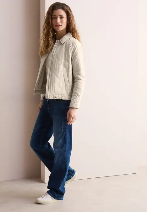 Frau mit heller Jacke, blauer Jeans und weißen Sneakern steht an heller Wand.