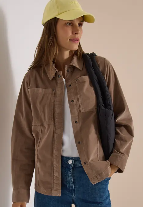 Eine Frau trägt eine beige Cordjacke, weißes Shirt, blaue Jeans, gelbe Kappe und schwarze Schultertasche.