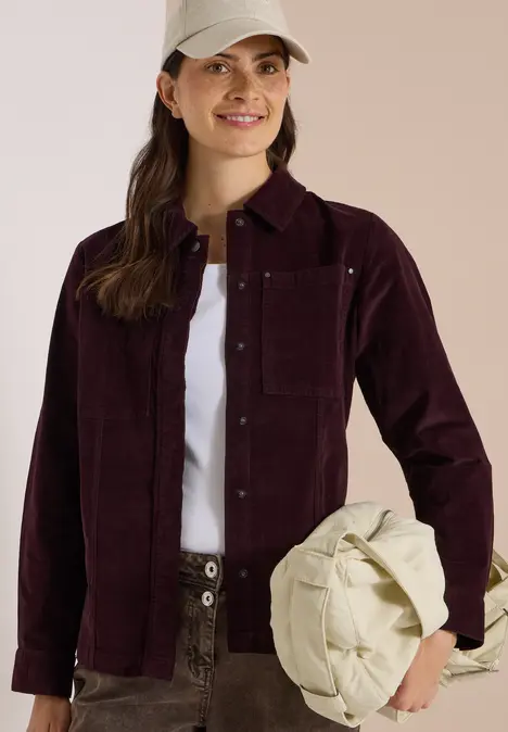 Frau in bordeauxfarbener Cordjacke, weißem T-Shirt und Cargohose hält lässig eine cremefarbene Tasche.