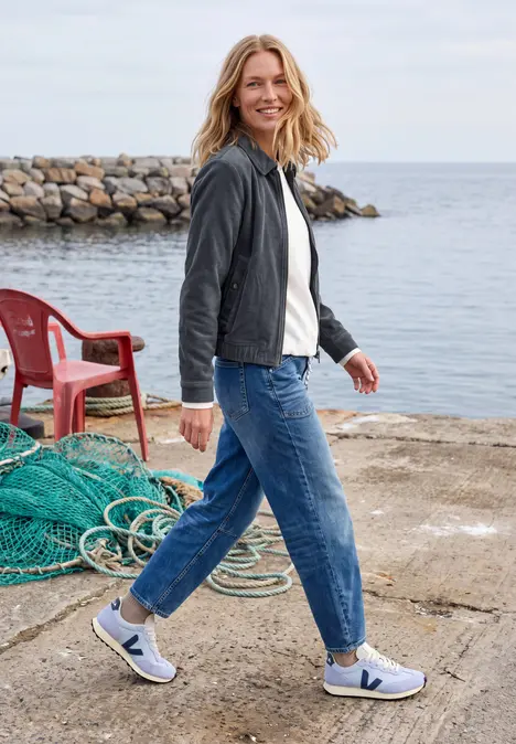 Eine Frau in Jeans und Sneakern läuft lächelnd auf einem Steg am Meer, im Hintergrund Fischernetz und Stuhl.