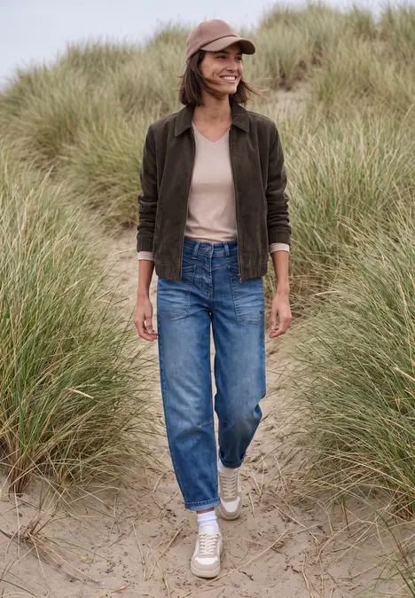 Eine Frau in Jeans, weißen Sneakers, brauner Jacke und Kappe spaziert lächelnd über Sanddünen.