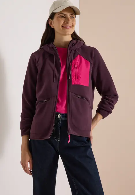 Frau trägt lila Kapuzenjacke mit pinker Brusttasche, Jeans und weiße Cap, lächelnd in Studio.
