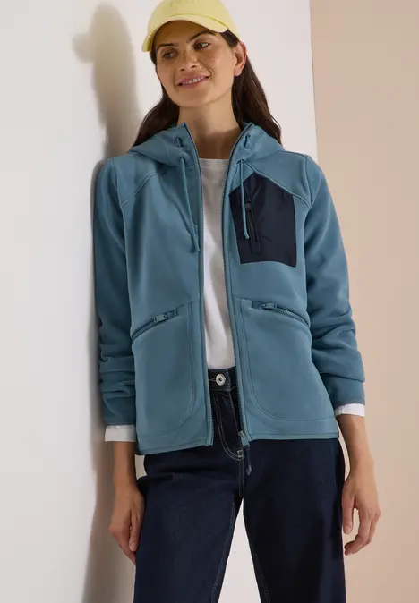 Frau in blauem Fleece-Hoodie, weißem Shirt, dunkler Hose und Cap vor neutralem Hintergrund.
