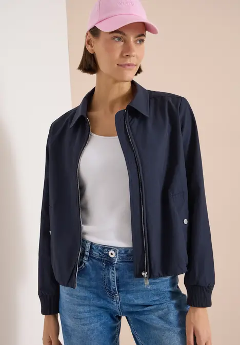 Eine Frau in Jeans, weißem Top, navyblauer Bomberjacke und rosa Cap, Gesicht verschwommen