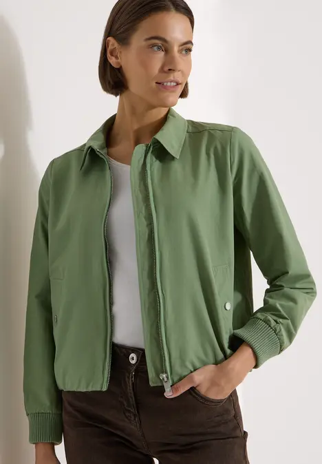 Person trägt grüne Hemdjacke, weißes Top und braune Hose in schlichtem, modischem Look.