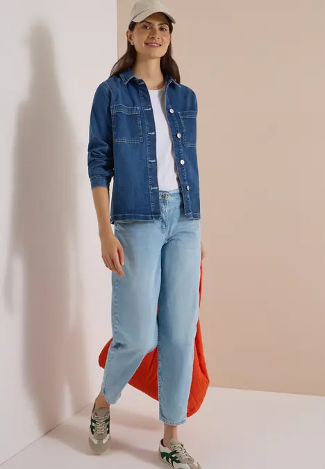 Frau in Jeansjacke, weißem T-Shirt, hellen Jeans und Sneakern trägt Cap und rote Tasche.
