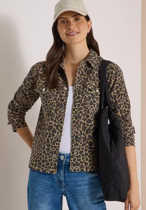 Frau in weißer Kappe, Leo-Bluse, Jeans und schwarzer Tasche lächelt locker.