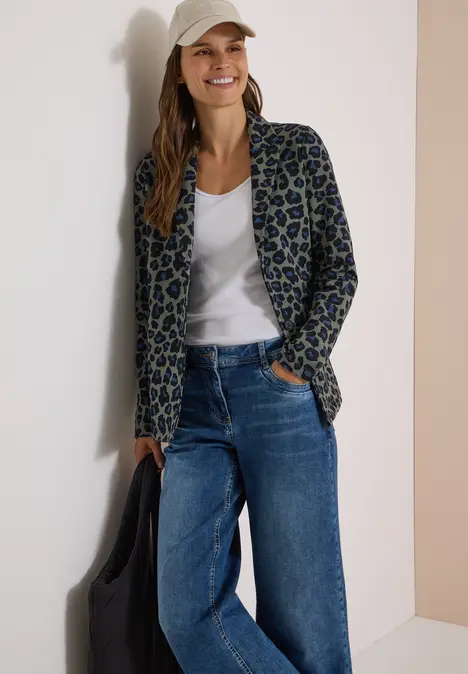 Frau lehnt entspannt an Wand, trägt Leopardenblazer, weißes Shirt, weite Jeans und Cap, lächelt.