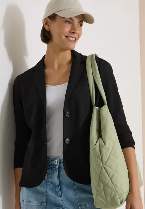 Person trägt schwarzen Blazer, weißes Top, Jeans und eine grüne, gesteppte Tasche über der Schulter.