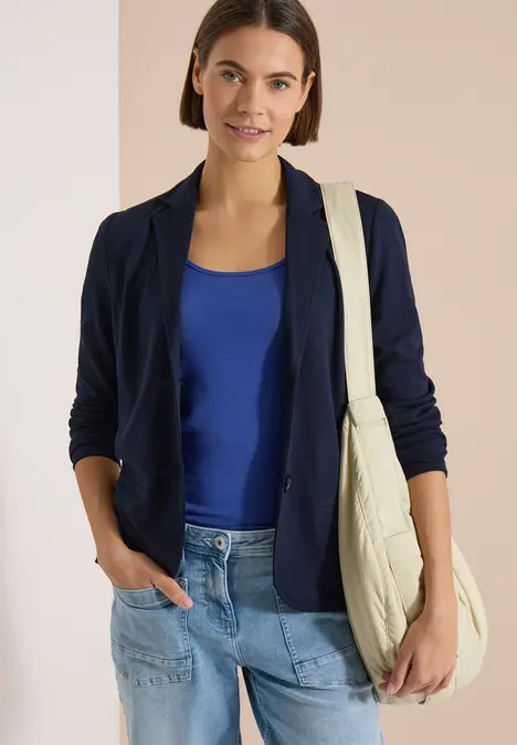 Frau in dunkelblauem Blazer, blauem Top und Jeans trägt eine beige Stepp-Tasche, lässig mit Hand in Tasche.