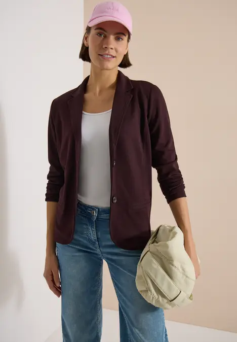 Person in Jeans, weißem Top, dunkelrotem Blazer und Cap hält eine hellbeige Tasche vor beigem Hintergrund.