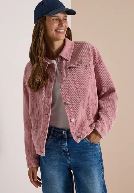 Junge Frau in rosafarbener Cordjacke, Jeans und blauer Cap lächelt entspannt.