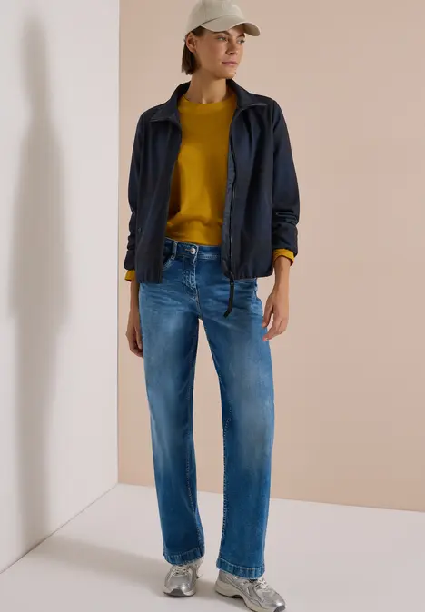 Frau trägt blaue Jeans, senfgelber Pullover, dunkelblaue Jacke und beige Mütze vor hellem Hintergrund.