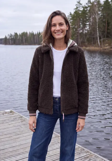 Frau lächelt auf Steg am ruhigen See, trägt dunkle Jacke, weißes Shirt und Jeans vor Waldkulisse.
