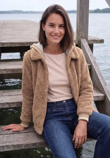 Junge Frau sitzt auf einer Holztreppe am Wasser, lächelt und trägt Jeans und eine kuschelige Jacke.