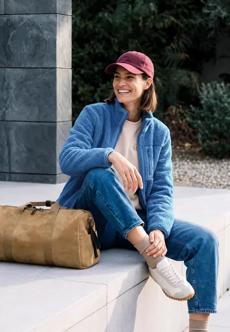 Eine Frau mit roter Mütze und blauer Fleecejacke sitzt lächelnd mit Tasche auf einer Steinbank.