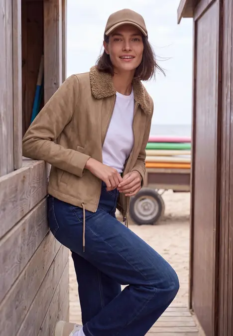 Frau in beiger Jacke, Jeans und Cap lehnt an Holzwand am Strand, im Hintergrund Sand und Meer.