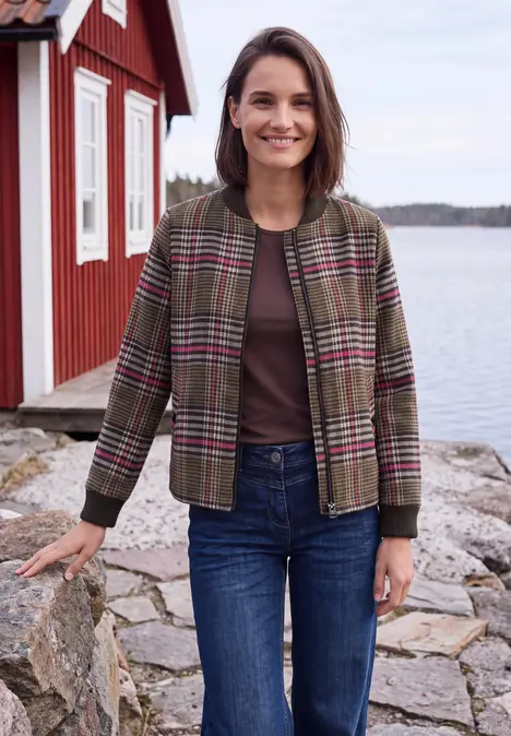 Lächelnde Frau in karierter Jacke und Jeans steht vor rotem Haus am Wasser.