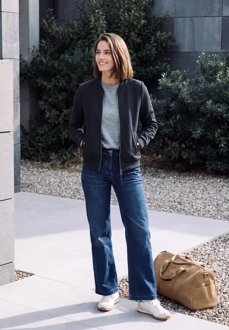 Eine Frau in Jeans und Jacke steht lässig mit Reisetasche vor einem modernen Gebäude und grünen Büschen.