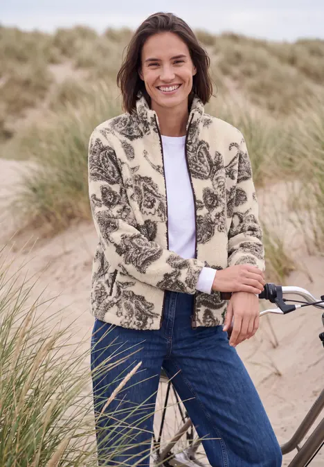 Frau in Blumenfleece und Jeans lächelt lässig neben Fahrrad am Dünenstrand.