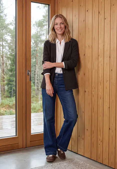 Eine lächelnde Frau in Jeans und Blazer steht vor einer Holzverkleidung neben großen Glastüren.
