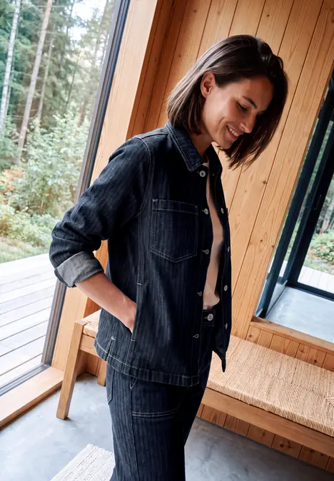 Frau im dunklen Denim-Outfit steht lächelnd am Fenster in einem hellen Holzraum.