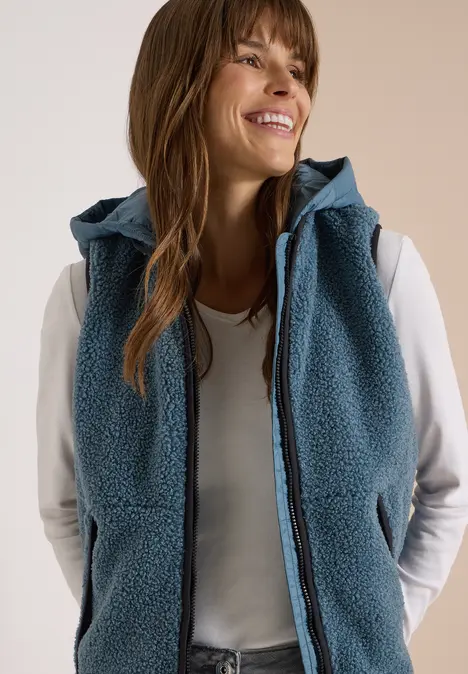Frau in weißem Langarmshirt und blauer, ärmelloser Fleece-Weste mit Kapuze, sie lächelt und schaut zur Seite.