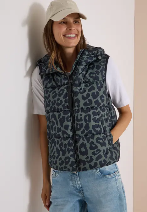 Junge Frau trägt Jeans, weißes T-Shirt, Leopard-Weste und Baseballcap, lächelt.