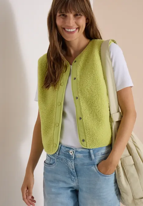 Frau mit grüner Strickweste, weißem T-Shirt und Jeans, lächelnd vor hellem Hintergrund.