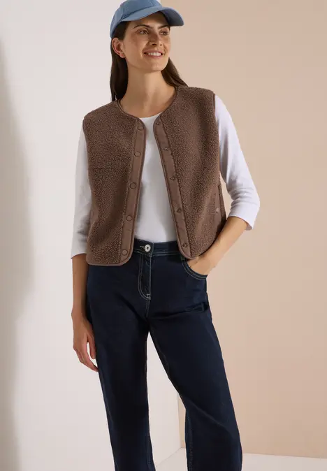 Eine junge Frau trägt eine braune Fleece-Weste, weißes Langarmshirt, blaue Jeans und eine blaue Baseballkappe.
