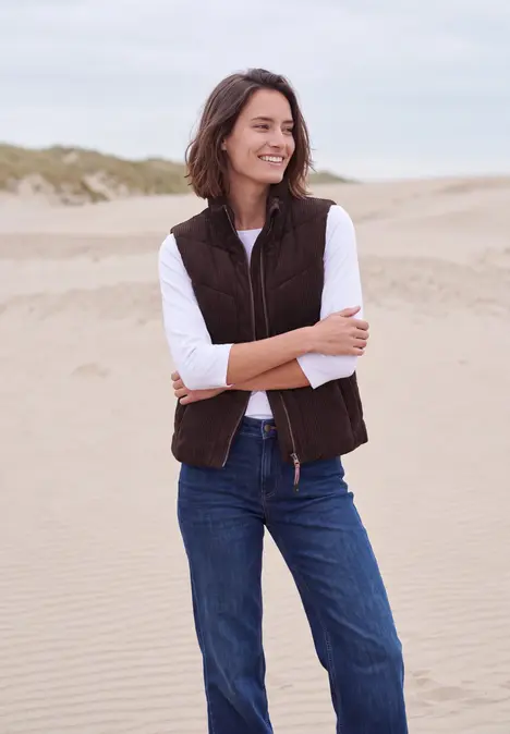 Eine Frau steht lächelnd am Strand in brauner Weste, weißem Shirt und blauen Jeans vor Sanddünen.