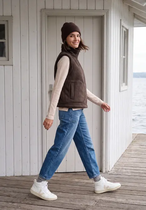 Eine junge Frau spaziert auf einem Holzsteg am See, trägt braune Weste, Beanie, Jeans und weiße Sneakers.
