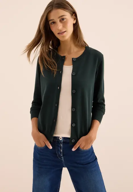 Frau in dunkelgrüner Strickjacke, weißem Shirt und Jeans vor hellem Hintergrund.