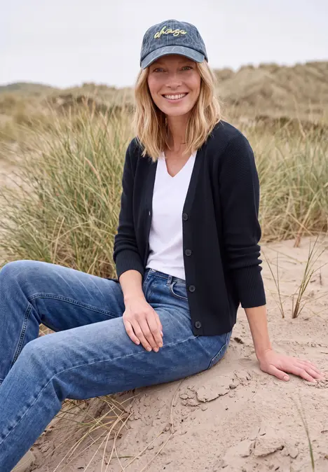 Eine Frau sitzt lächelnd in den Dünen, trägt Jeans, weißes T-Shirt, schwarze Strickjacke und Cap.