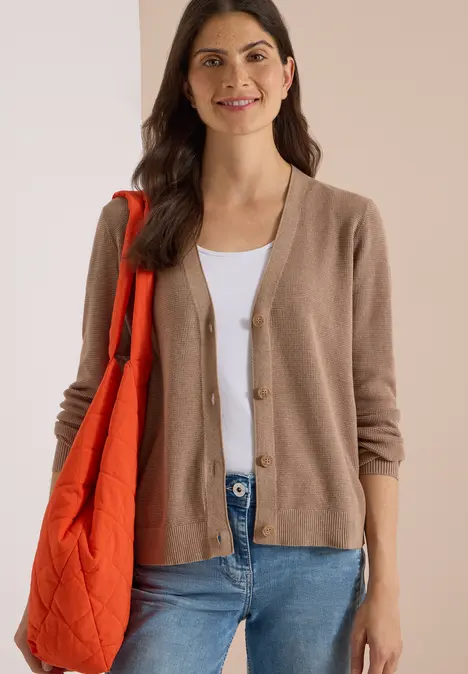 Braunhaarige Frau mit beigefarbener Strickjacke, weißem Top, blauen Jeans und oranger Umhängetasche lächelnd.