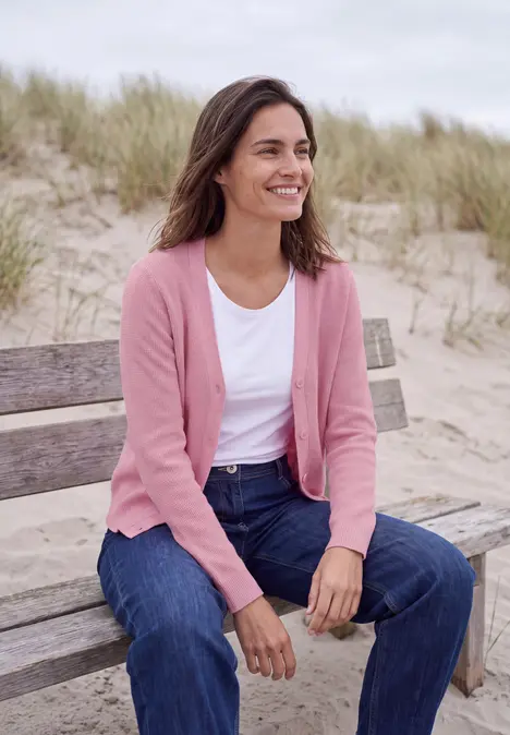 Kobieta uśmiechnięta w różowym kardiganie siedzi na ławce plażowej, za nią trawy wydm.