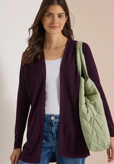 Frau in dunkelviolettem Cardigan, weißem Top, Jeans, mit großer, grüner Stepp-Tasche über der Schulter.