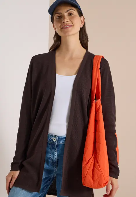 Frau in Jeans, weißem Top, dunkler Strickjacke, blauer Mütze und orangefarbener Tasche