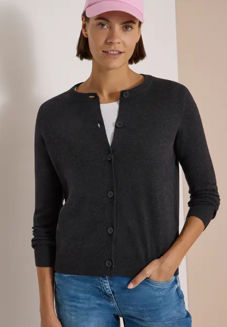 Frau in grauer Strickjacke, Jeans und rosa Cap vor schlichtem zweifarbigem Hintergrund.