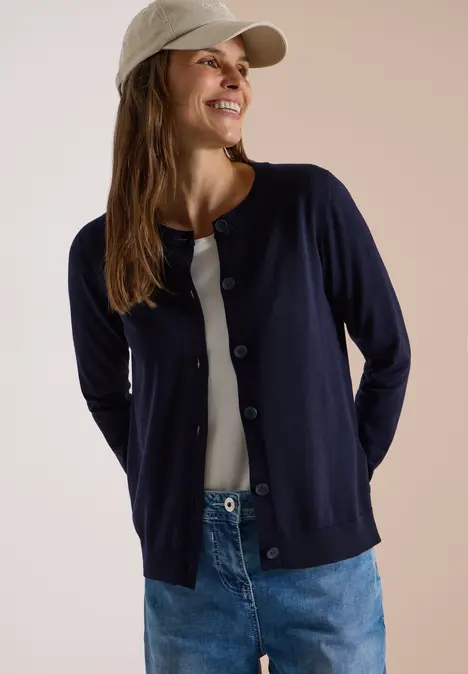 Frau in Jeans, weißem Top, dunkelblauer Strickjacke und Cap lächelt vor hellem Hintergrund.
