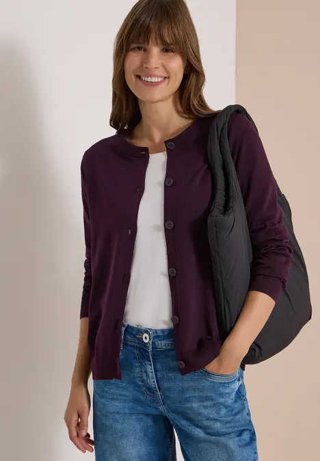 Eine lächelnde Frau trägt eine lila Strickjacke, weißes Shirt, blaue Jeans und eine schwarze Schultertasche.