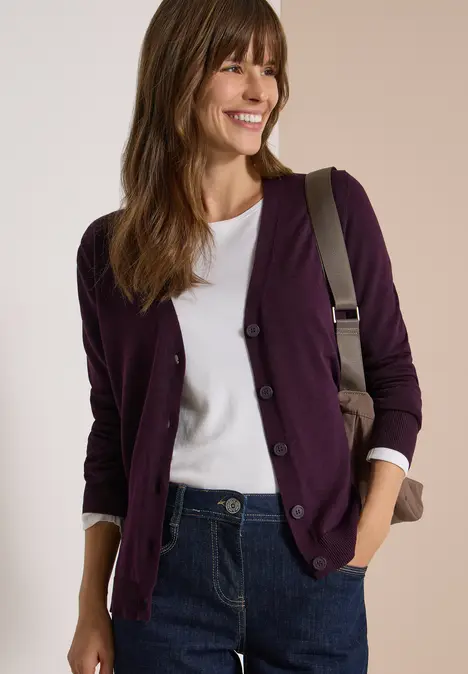 Eine lächelnde Frau trägt eine lila Strickjacke, weißes Shirt, Jeans und Umhängetasche.