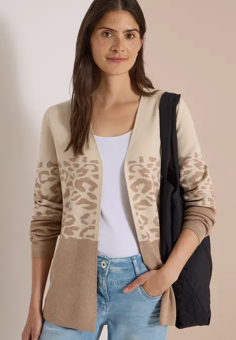 Frau in weißem Top, beiger Strickjacke mit Animalprint und schwarzer Tasche vor neutralem Hintergrund.