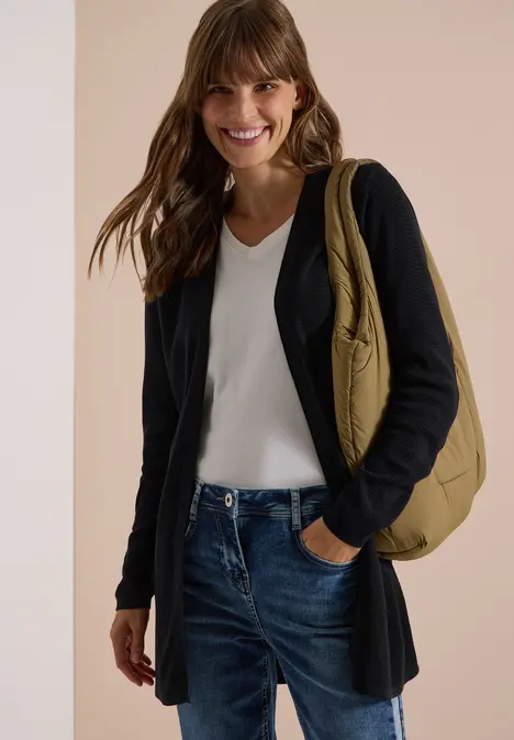 Frau in dunkler Strickjacke, weißem Top und Jeans, lächelt, trägt eine bequeme Umhängetasche.