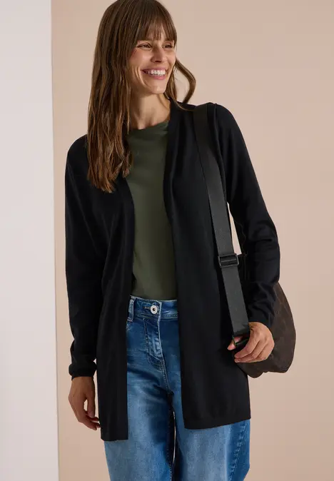 Eine Frau trägt einen schwarzen Cardigan, grünes Shirt, Jeans und lächelt, mit Tasche über der Schulter.