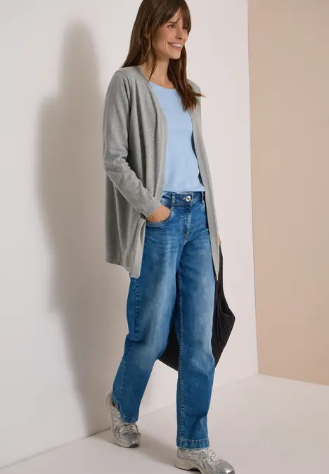 Frau trägt graue Strickjacke, hellblaues Top, weite Jeans und Sneaker, hält eine schwarze Tasche.
